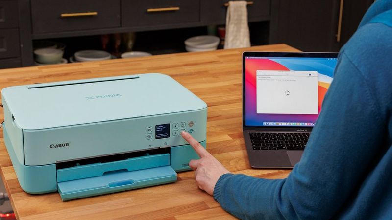 A woman is pressing an OK button on the Canon printer, laptop is seen in the background with Wi-Fi Connection Assistant software on the screen.