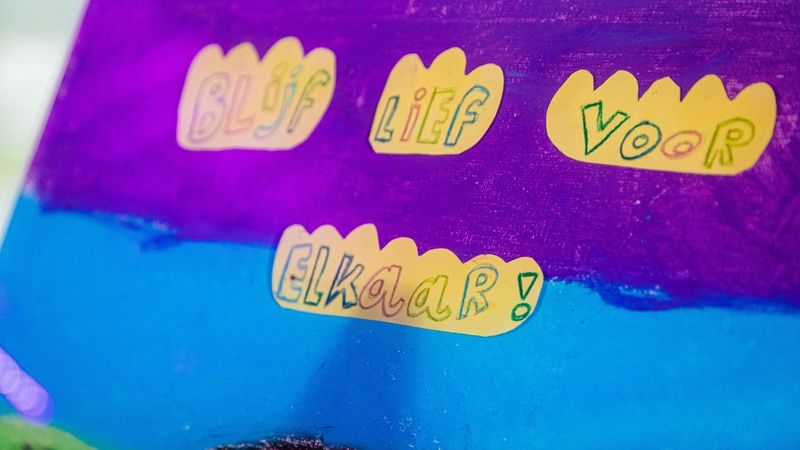 A childs handwriting on a painted purple and blue background. In Dutch it reads, Blijf life voor elkaar!, which in English means Always be kind to each other!.