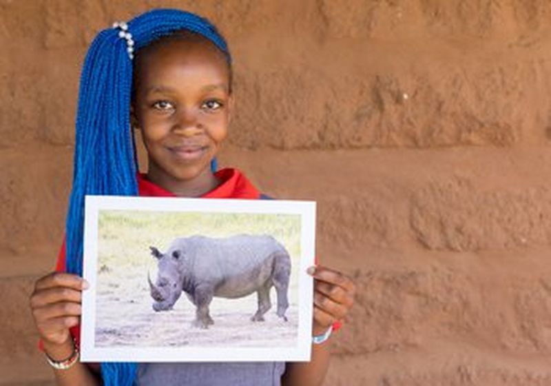 A child with long bright blue braids and a red t-shirt holds up a photograph of a rhino. She stands against a terracotta-coloured wall.