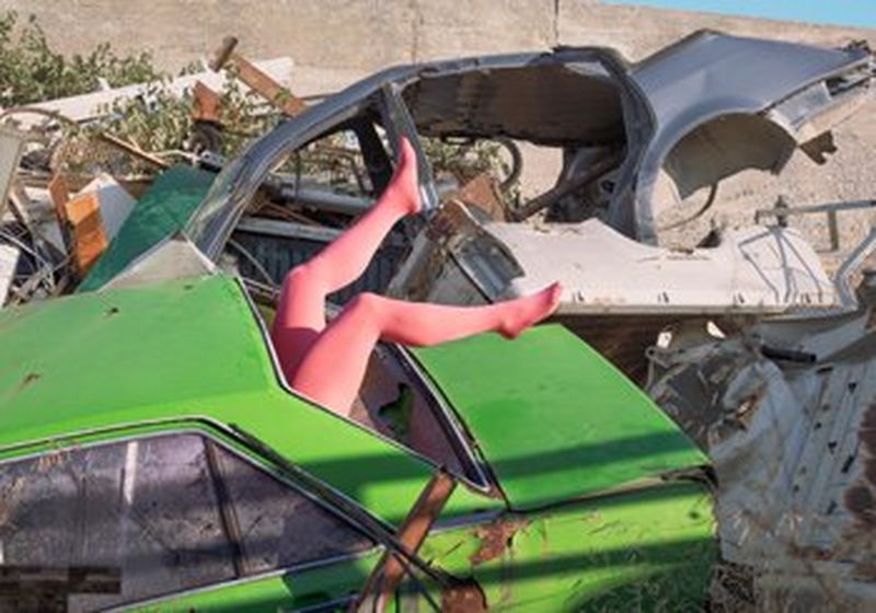 A bright green car lies discarded at an angle in a junk yard, surrounded by other discarded objects and car parts. The rear window of the car is gone and a pair of what appear to be women’s legs in pink tights protrude through where the window should be. It is impossible to tell if they are human or a mannequin.