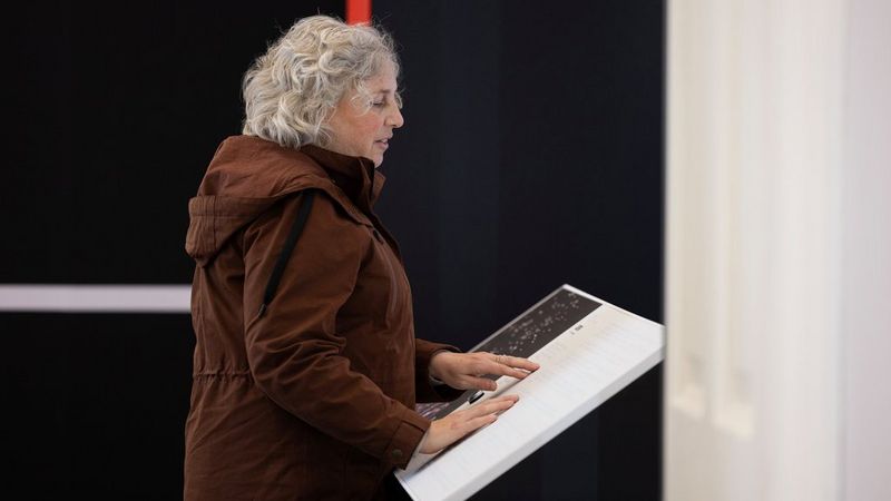 A visually impaired woman in a brown jacket with short, grey curly hair uses her hands to read the braille content of an exhibition information plinth.