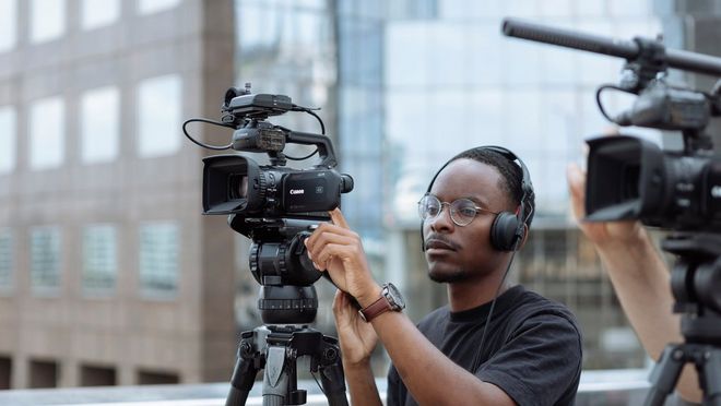 Een persoon filmt met een Canon XA75-videocamera op een statief, met grote kantoorgebouwen op de achtergrond.