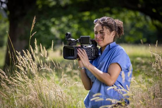 A woman wearing a blue shirt films with a Canon XF605 camcorder in a field of tall grass.