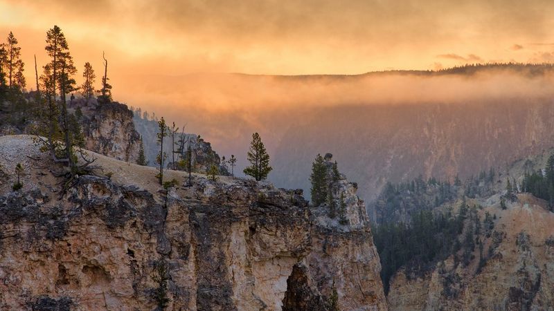 A dramatic landscape photo of a canyon at sunset. Orange and pink hues fill the sky, casting a warm glow on the rocky cliffs and pine trees below. Mist hangs low in the valley, creating a mysterious atmosphere.