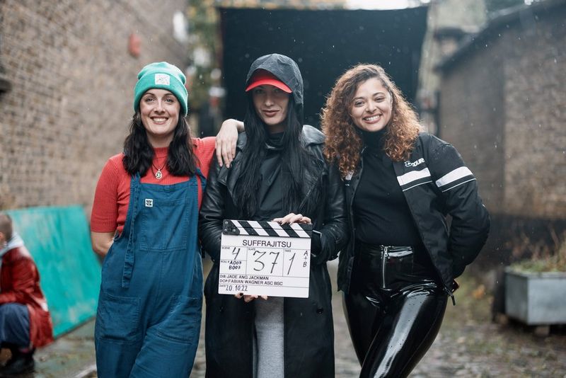 Three young women stand close together, smiling at the camera. There is a cobbled and brickwork street behind them, and the middle woman is holding a clapperboard labelled "SUFFRAJITSU".
