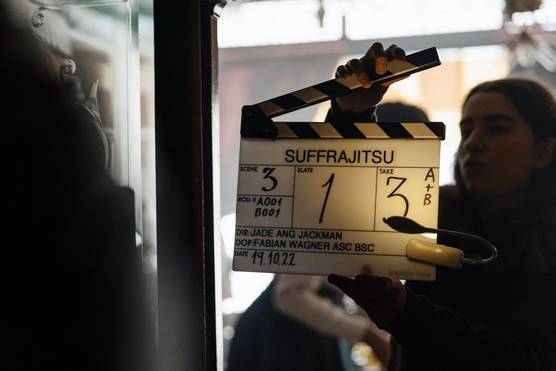 A woman holds up a clapperboard labelled "SUFFRAJITSU", and showing Scene 3, Slate 1 and Take 3.