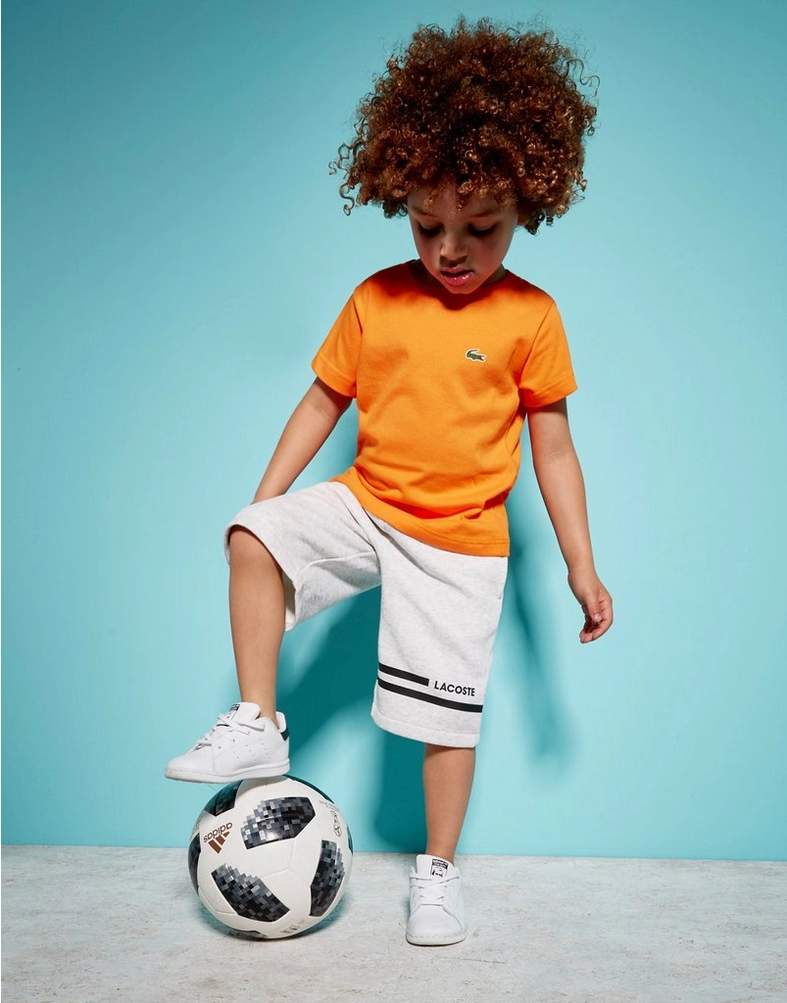 På billedet ses en lille dreng, som er klædt i Lacoste tøj. Drengen har en orange t-shirt på med Lacostes verdenskendte krokodille på brystet, og et par hvide fleece shorts fra Lacoste. Drengen står og leger med en fodbold.