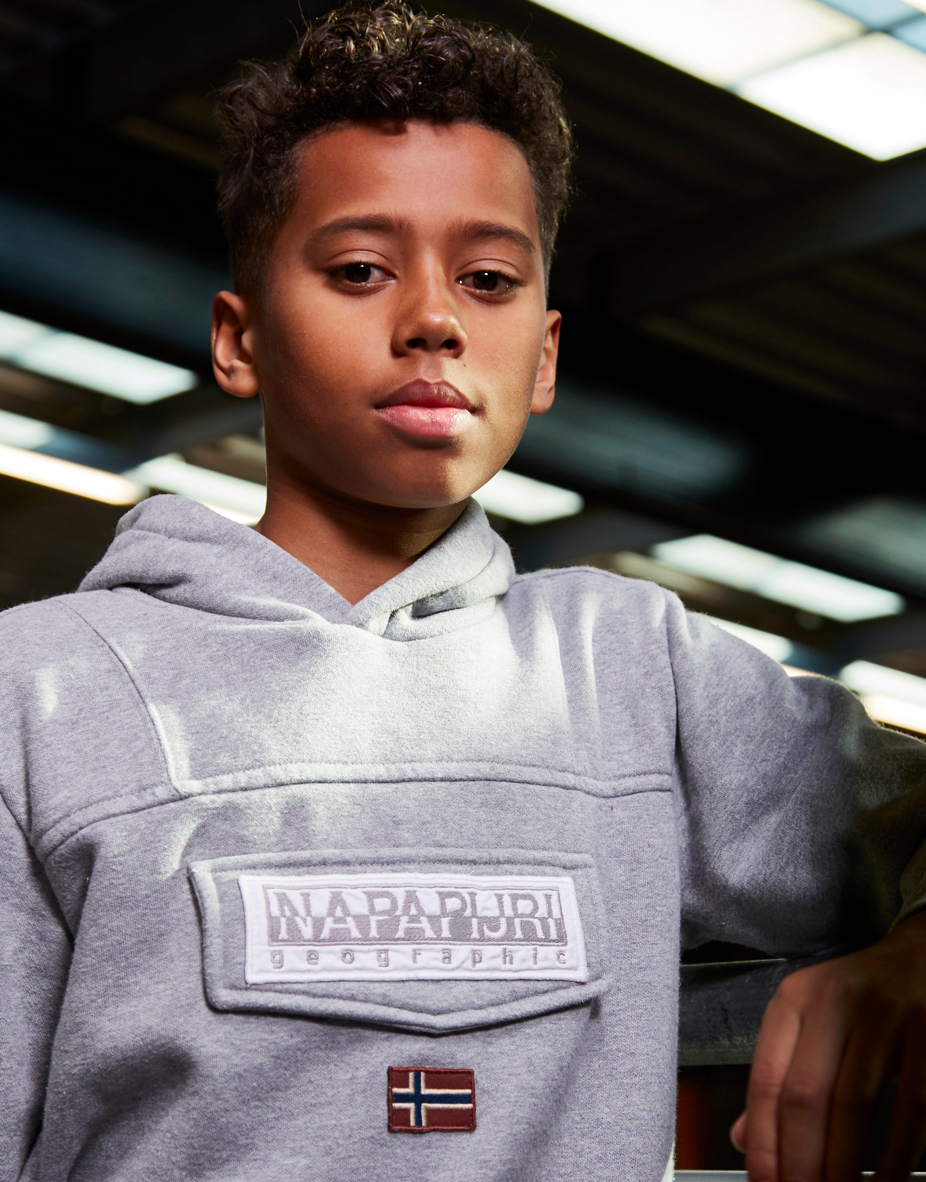 På billedet ses en ung dreng, som er klædt i en trøje fra Napapijri. Trøjen er grå og udstyret med hætte og kængurulomme på maven. 