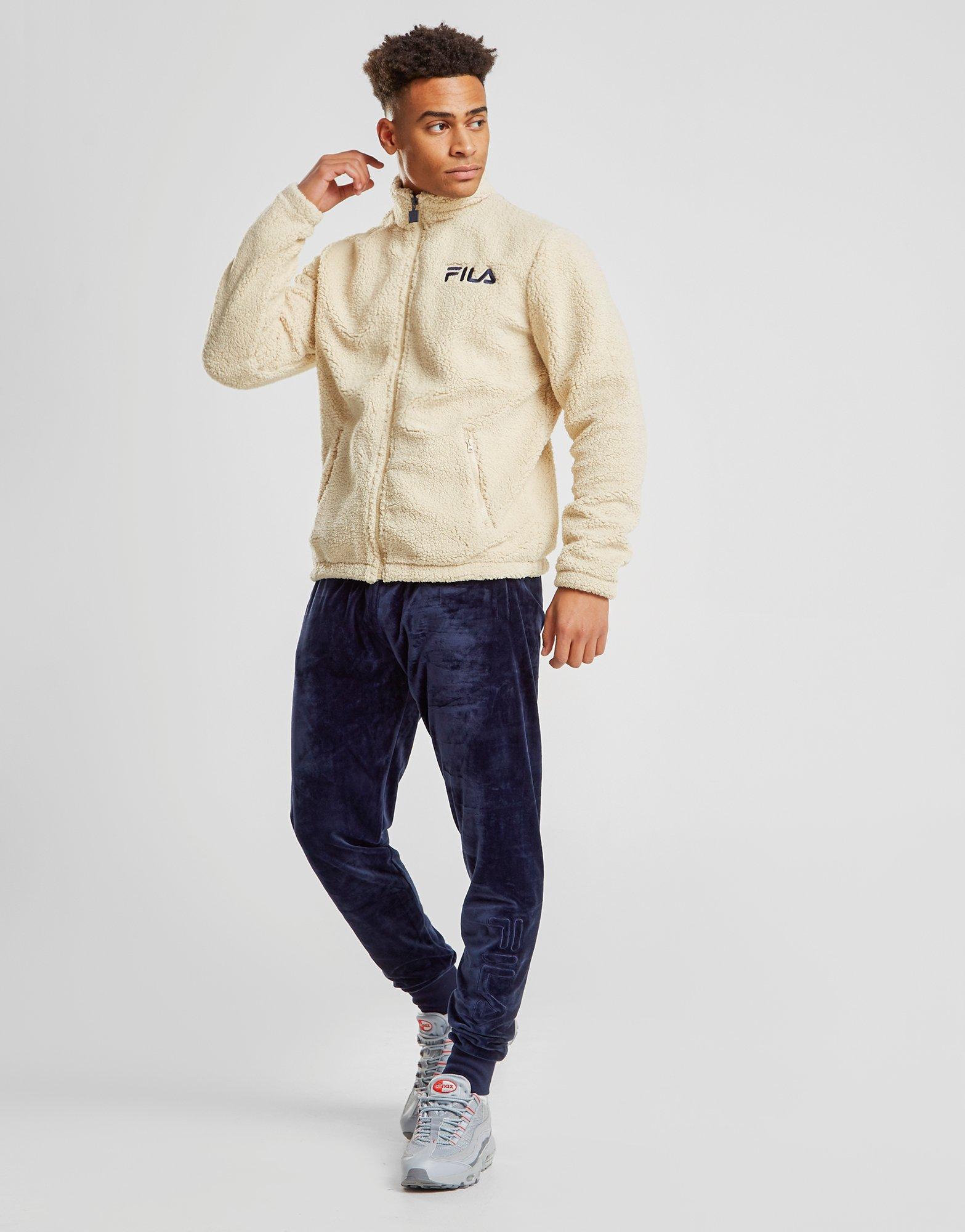 På billedet ses en ung mand, som er klædt i Fila tøj. Jakken er en Fila Gordon Borg Fleece Jakke i hvid/beige, og bukserne er Fila Aaron Velour Bukser i blå. Fås eksklusivt hos JD Sports!